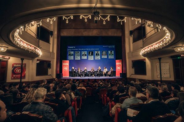 Panel of eight speakers on stage at a conference inside a vintage theater with a full audience and chandeliers overhead.