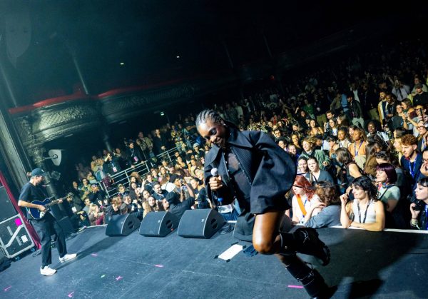 Performer in a black outfit holding a microphone dancing on stage while a guitarist plays and a large crowd watches.