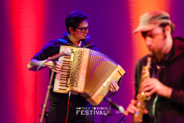 A musician playing an accordion and another playing a saxophone on stage with a red and purple lit background, at a festival named 