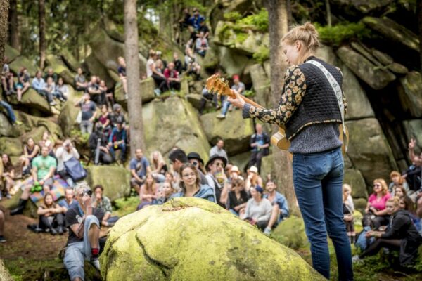 A woman playing a guitar to an audience seated on rocky terrain in a forested area.