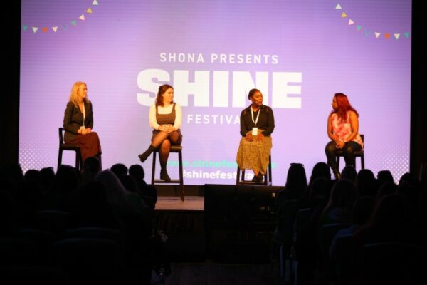 Four women participating in a panel discussion on stage at the SHINE Festival, with the audience in the foreground and a banner in the background displaying the event's name and website.