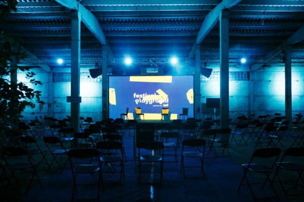 An empty event space with rows of chairs facing a large screen displaying the words 'festival playground', illuminated in blue light with shadows of plants on the side walls.