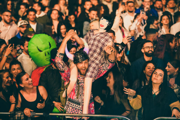 A crowd of people at a concert or event with some wearing costumes and masks, one person is holding up a plastic doll mannequin, and many in the audience are taking photos or recording the event.