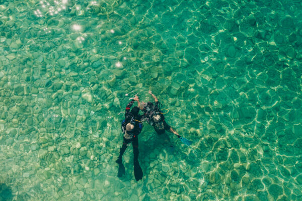 Aerial view of two scuba divers in clear, shallow water over a rocky bottom with sunlight reflecting on the surface.