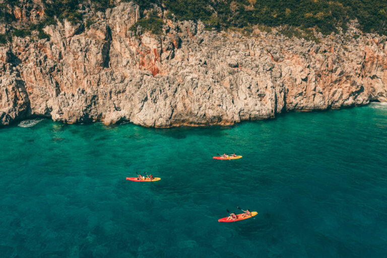 Three kayakers on a turquoise sea near a rugged cliff coastline.