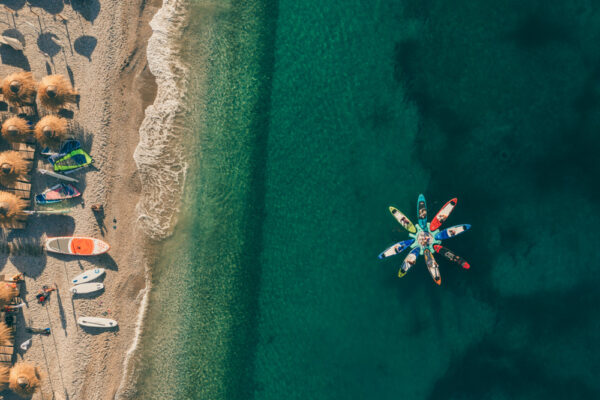 Aerial view of a sandy beach with straw umbrellas and water sports equipment, next to a clear sea with a group of people on colorful kayaks arranged in a circular 'flower' pattern.