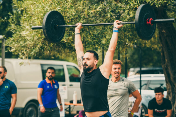 A man in a black shirt with a full beard and a weightlifting belt lifting a barbell overhead with outstretched arms, as onlookers watch in the background outdoors.