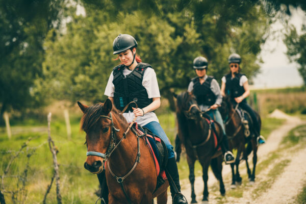 Three people wearing protective riding gear, including helmets and safety vests, are horseback riding along a rural trail, with the focus on the smiling young rider in the foreground.