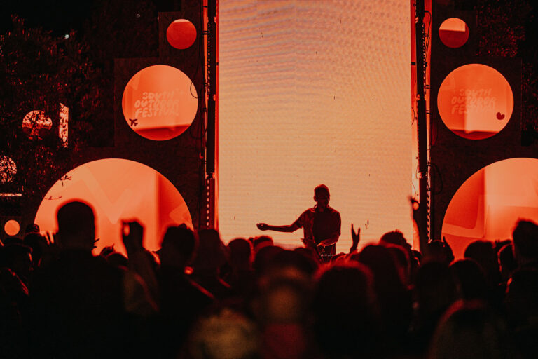 Silhouette of a DJ performing at a music festival with enthusiastic crowd in the foreground, large illuminated screen and festival branding in the background. The scene is bathed in a warm red light.
