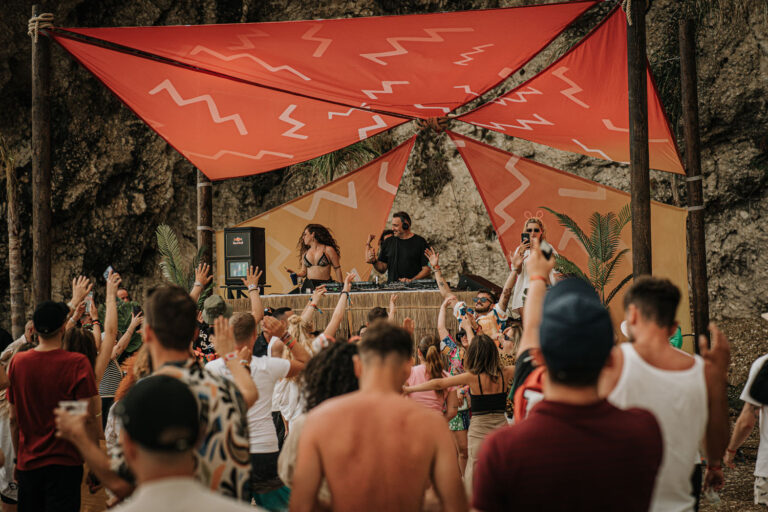 A vibrant outdoor DJ event with a lively crowd dancing and raising their hands under a red canopy, with a female DJ performing alongside a male DJ, surrounded by lush greenery and rocky formations.