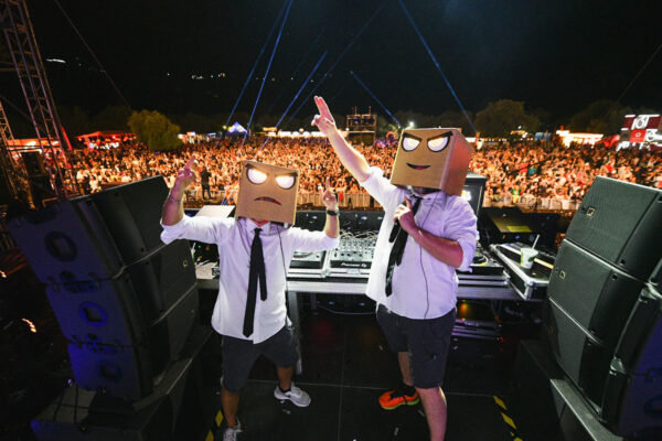 Two performers with cardboard box heads gesturing to the crowd at a night-time outdoor music festival.