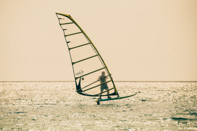 A windsurfer gliding over the water at sunset, with the sunlight reflecting off the waves and the sail silhouette against a warm, glowing sky.