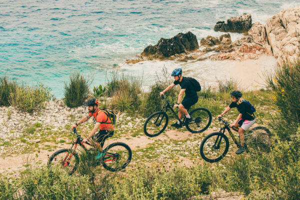 Three cyclists riding mountain bikes on a coastal trail with the sea and rocky shoreline in the background.