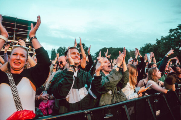 A crowd of enthusiastic concert-goers cheering and clapping at an outdoor music festival with some people making heart shapes with their hands.