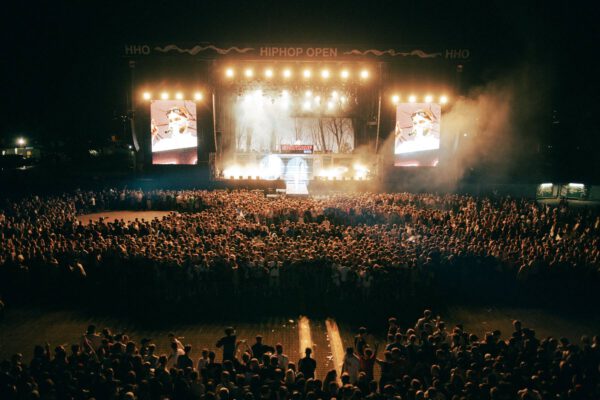 A large crowd at an outdoor hip-hop concert at night with stage lights and large screens displaying a performer.