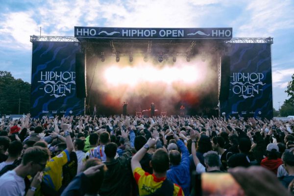 A large crowd of people at an outdoor Hip Hop Open music festival with hands raised enjoying the performance, with the stage in the background, colorful stage lighting, and banners displaying the event name.