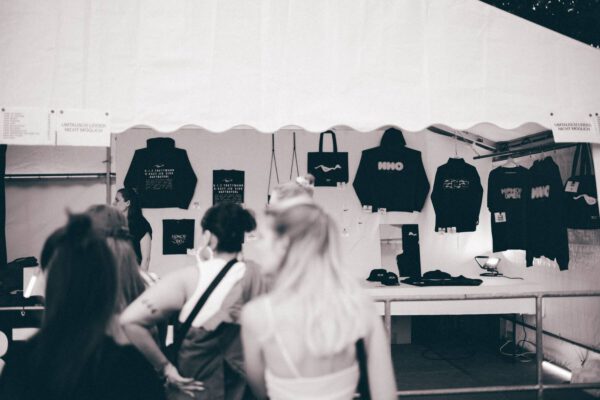 A monochrome image of potential customers at a merchandise stall with various black clothing items and accessories displayed, including t-shirts and bags with white text and graphics.
