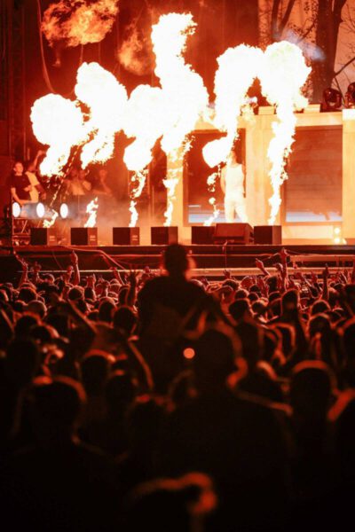 A concert stage with intense flames shooting up, illuminating the performers and a lively crowd in the foreground.
