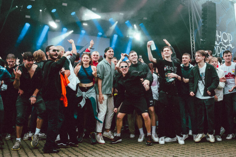 A group of enthusiastic people enjoying themselves at a music festival with some of them raising their arms, smiling, and posing for the photo. The stage lights cast beams across the scene, enhancing the festive atmosphere.
