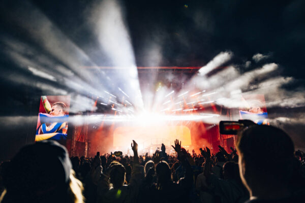 A lively concert crowd with raised hands, silhouetted against a brightly lit stage with beams of light and smoke effects, with a large screen displaying a performer.