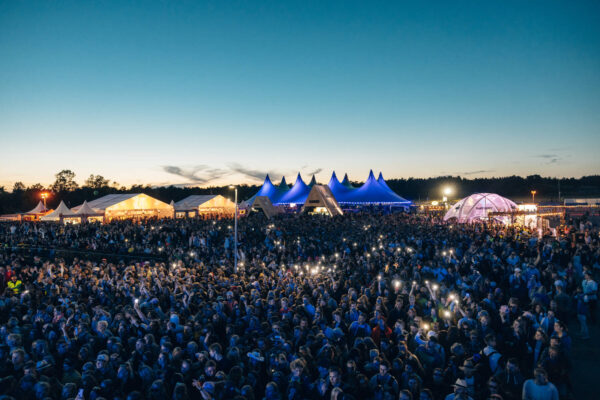 A large outdoor festival crowd at dusk with multiple illuminated mobile phone screens, set against a backdrop of distinctive blue-topped tents and a softly glowing stage in a rural setting.
