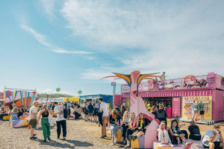 A lively outdoor festival scene with people milling around colorful food stalls, seating areas, and a pink double-decker bus structure under a clear blue sky.