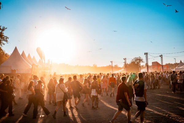 A bustling outdoor festival scene with crowds of people walking and the sun setting in the background, creating a hazy, golden atmosphere, with birds flying overhead and festival tents and banners visible along the sides.