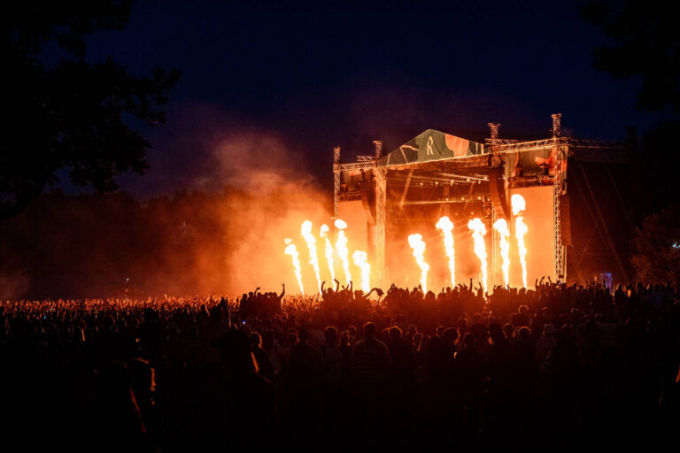 A large crowd gathered at a night-time outdoor concert with bright stage pyrotechnics illuminating the scene.