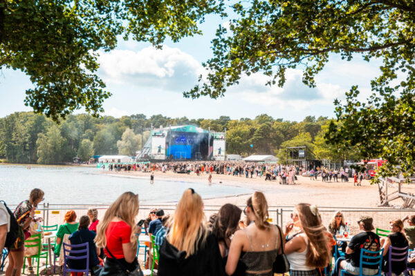 A vibrant outdoor music festival by a lake with a crowd of people standing and sitting on the sandy shore, with a stage in the distance framed by trees under a clear sky.
