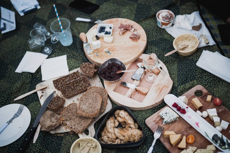 A picnic spread featuring various cheeses on wooden boards, sliced bread, bowls of dips, glasses of beverages, and utensils, arranged on a patterned blanket.