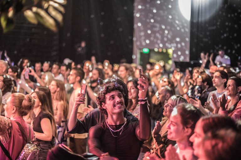 A crowd of people at a concert cheering and taking pictures with their phones, with bokeh lights in the background, and a featured man in the foreground looking up and smiling while taking a selfie.