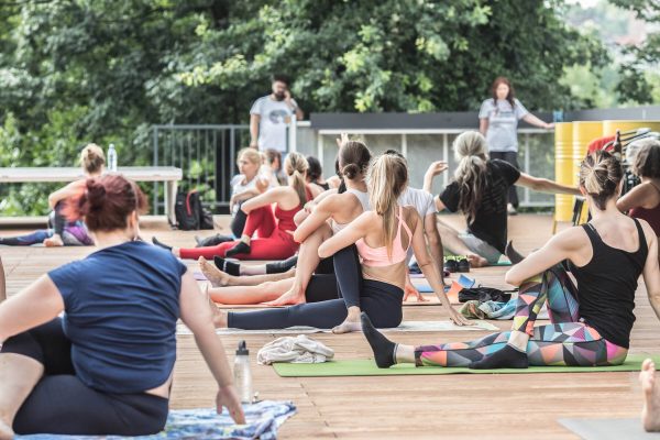 Group of people participating in an outdoor yoga class on a wooden deck surrounded by greenery, with a female instructor standing in the background.