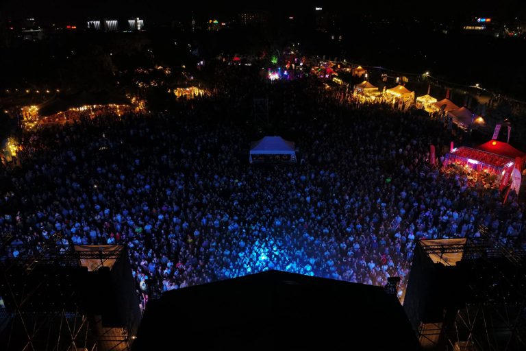 Aerial view of a crowded outdoor music festival at night, with colorful stage lights illuminating the audience and tents scattered around the venue.