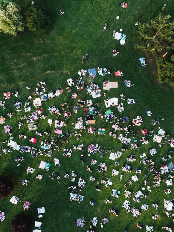 Aerial view of a public park with people sitting on various picnic blankets scattered across a grassy area with trees around the edge.