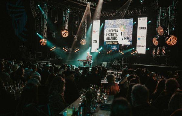 A large audience seated at round tables watches a speaker at a podium on stage during the European Festival Awards 2022 event, with banners and stage lighting around the venue.