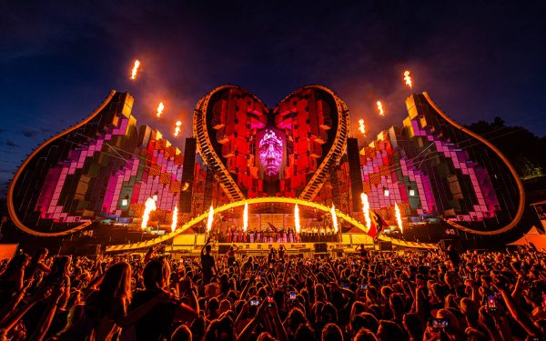 An elaborate outdoor music festival stage at night with a large heart-shaped LED screen backdrop, pyrotechnics shooting flames into the air, and a crowd of people with their hands up, some taking photos with smartphones.
