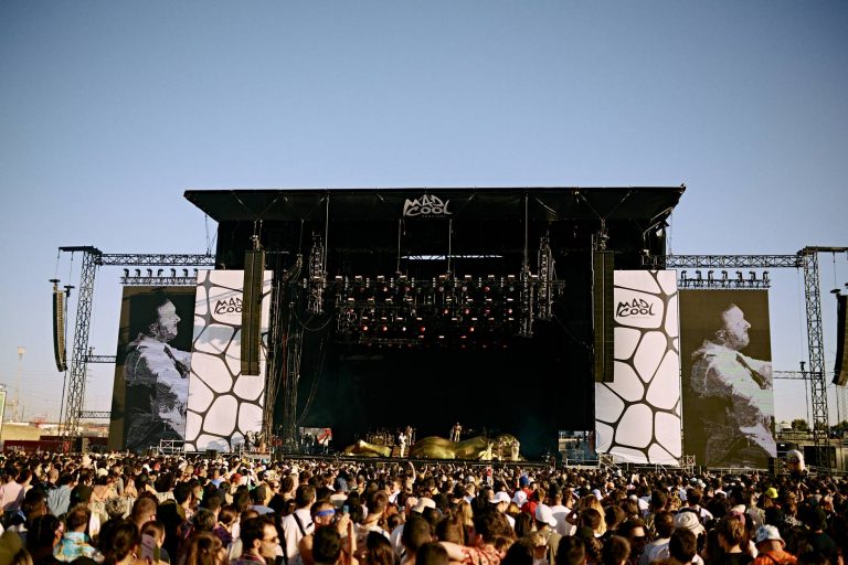 A large crowd of festival-goers enjoying a live performance at the Mad Cool Festival with the stage showing a band performing and two large screens displaying a musician.