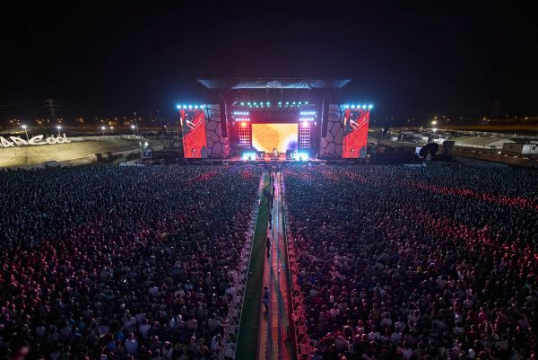 A large outdoor concert with a vast crowd gathered in front of a brightly lit stage at night, featuring prominent stage screens and lighting.