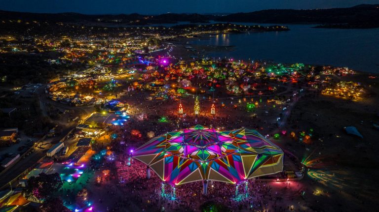 Aerial night view of a vibrant music festival with colorful lights, large tents, and a bustling crowd, set near a body of water with a backdrop of hills.