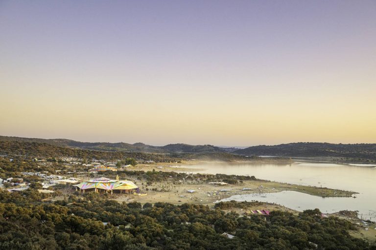 Aerial view of a colorful outdoor festival by a lake at dusk, surrounded by rolling hills and trees.
