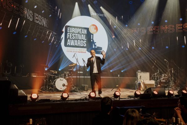 A man presents an award at the European Festival Awards 2023 event, standing on a stage with musical instruments and lighting equipment, under a spotlight and in front of a seated audience.