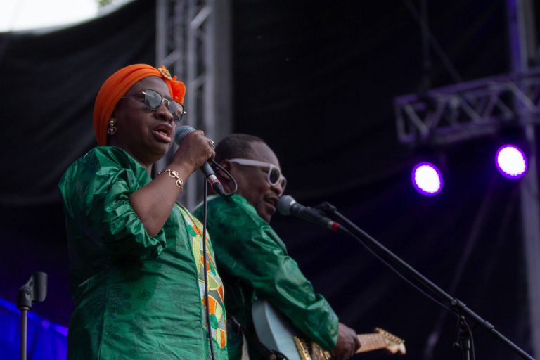 A woman wearing an orange headscarf and green outfit singing into a microphone with a man in sunglasses playing the guitar in the background on a stage with purple stage lights.