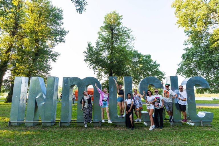 A group of people posing and smiling in front of large, stand-alone letters that spell out "MUSIC" in a park with trees in the background.