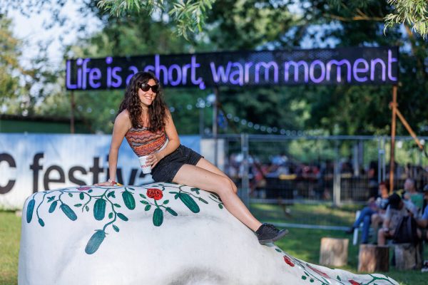 A smiling woman wearing sunglasses sits atop a white sculpture with painted green cucumbers and red flowers in an outdoor festival setting, with the blurry background featuring a sign that says 