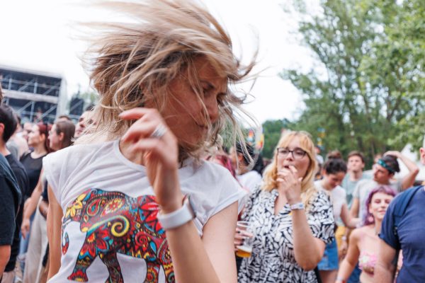 A woman in focus dancing with her hair flying, wearing a colorful elephant t-shirt, at a blurred outdoor music festival with other attendees in the background.