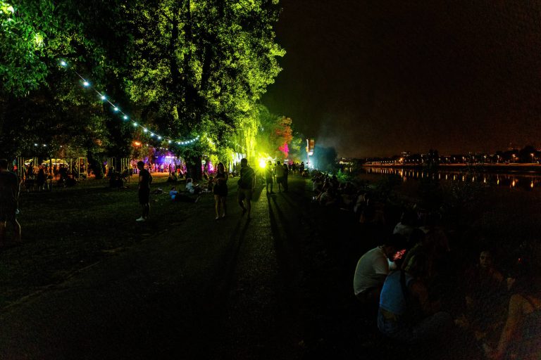 A nighttime scene of a lively outdoor event by a body of water, featuring people seated on the grass, walking, and socializing under string lights, with colorful stage lights visible in the distance and a city skyline barely illuminated along the horizon.