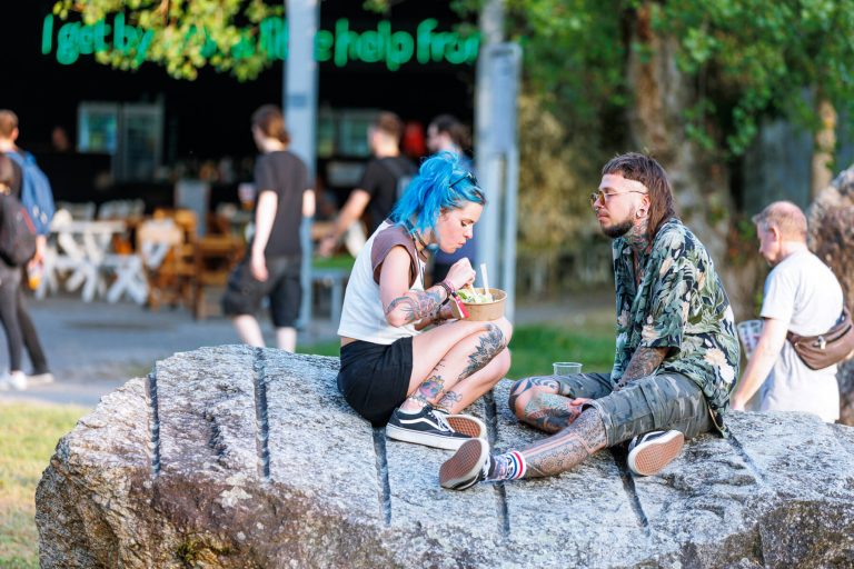 Two people with colorful hair and tattoos are sitting on a large rock, eating and conversing, with an outdoor food stand and other park-goers in the blurry background.