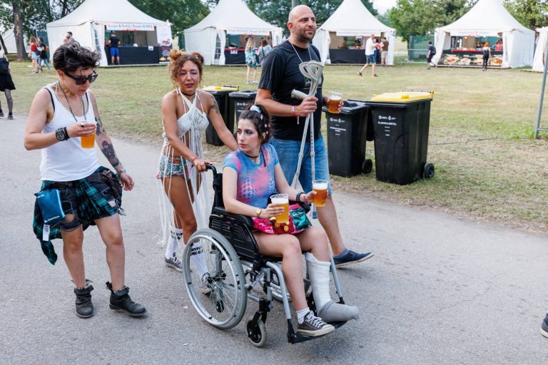 Four individuals at an outdoor festival with two people walking, one person in a wheelchair, and one person on crutches, all holding drinks.