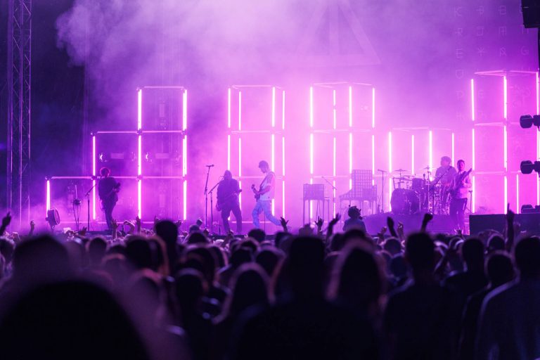 A live concert scene with a band performing on stage, illuminated by pink and blue stage lights, with silhouettes of the audience in the foreground.