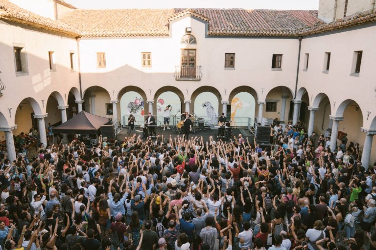 Outdoor concert with a crowd of people raising their hands towards a band performing in a courtyard with arches and murals.